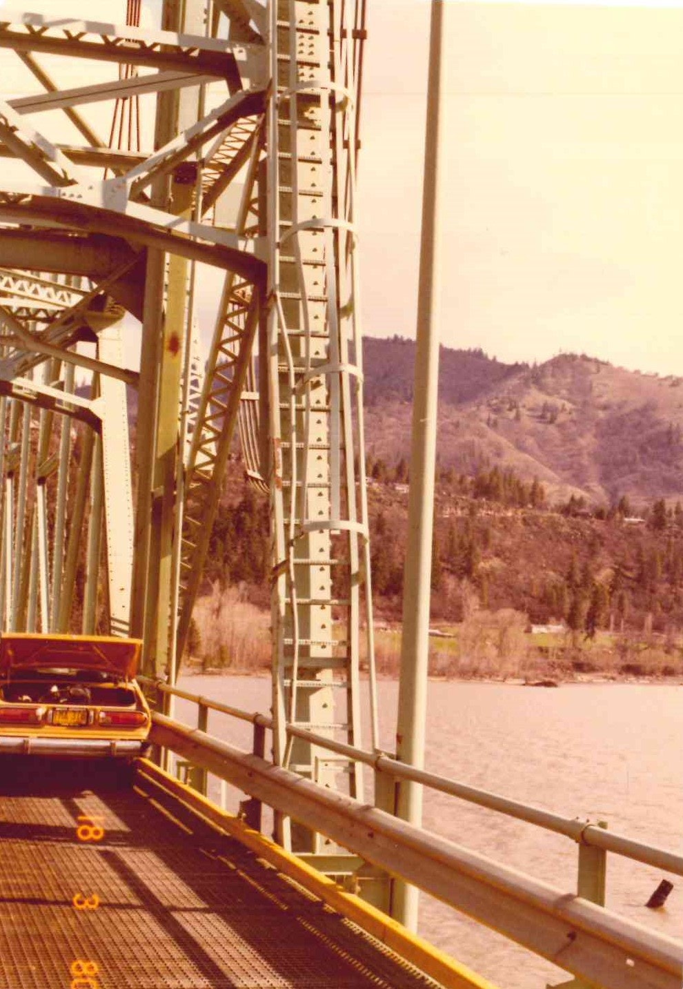 Vintage photo of a car crossing a steel truss bridge over a river with hills in the background.