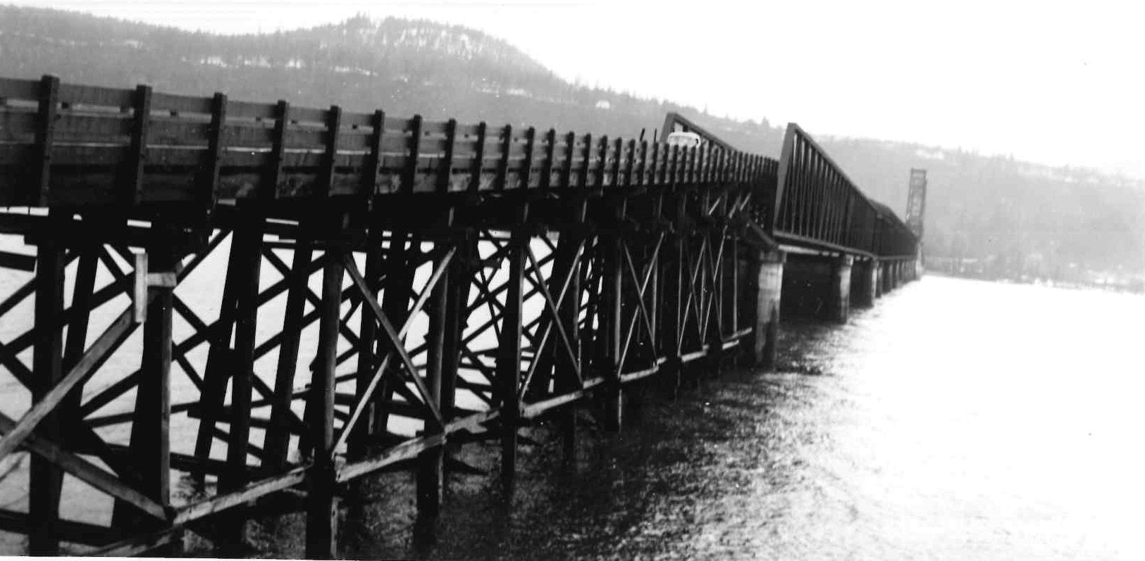 Black and white photo of a covered wooden trestle bridge over water with hills in the background.