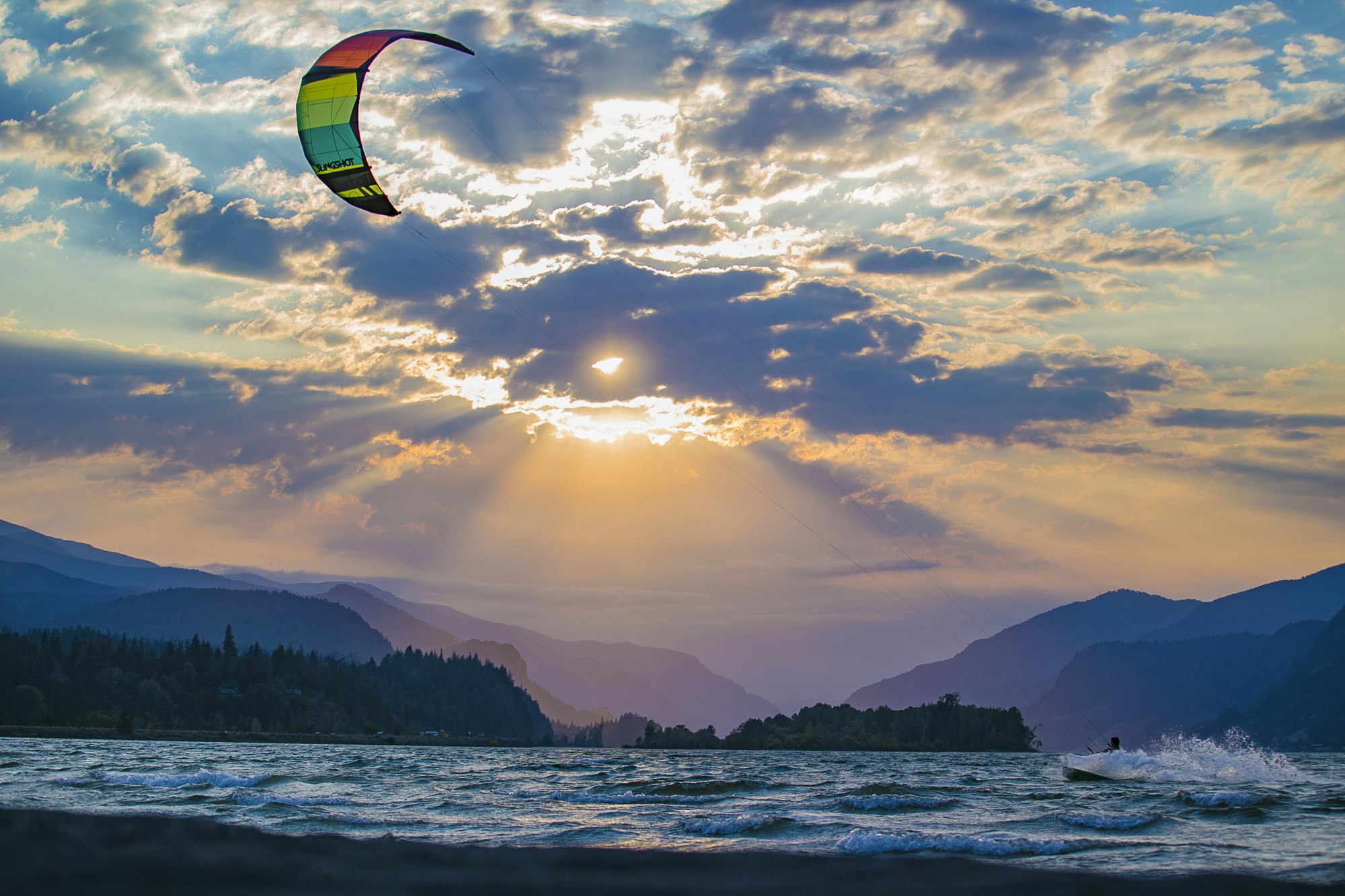 A kite surfer on choppy waters at sunset, with rays of sunlight beaming through cloud cover over mountains.