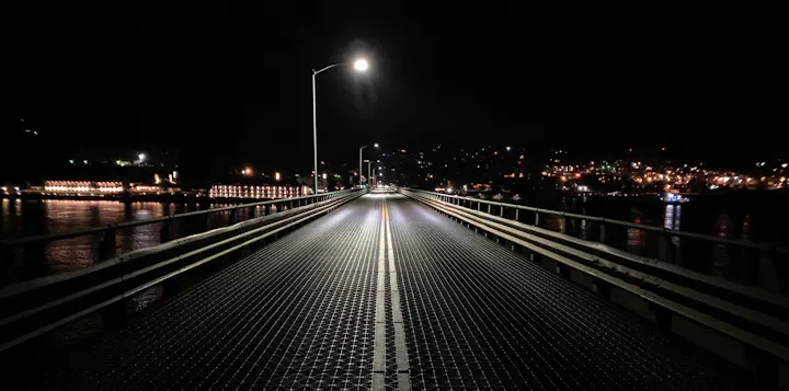 A dimly lit bridge at night, with city lights reflecting on the water, creating a serene and atmospheric scene.