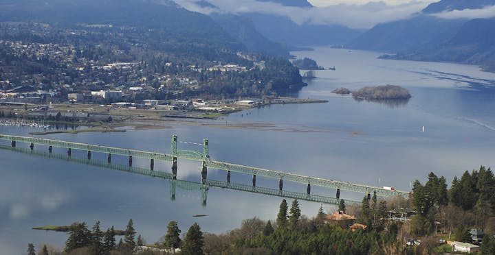 A scenic view of a green bridge over a wide river, with mountains and a town in the background.
