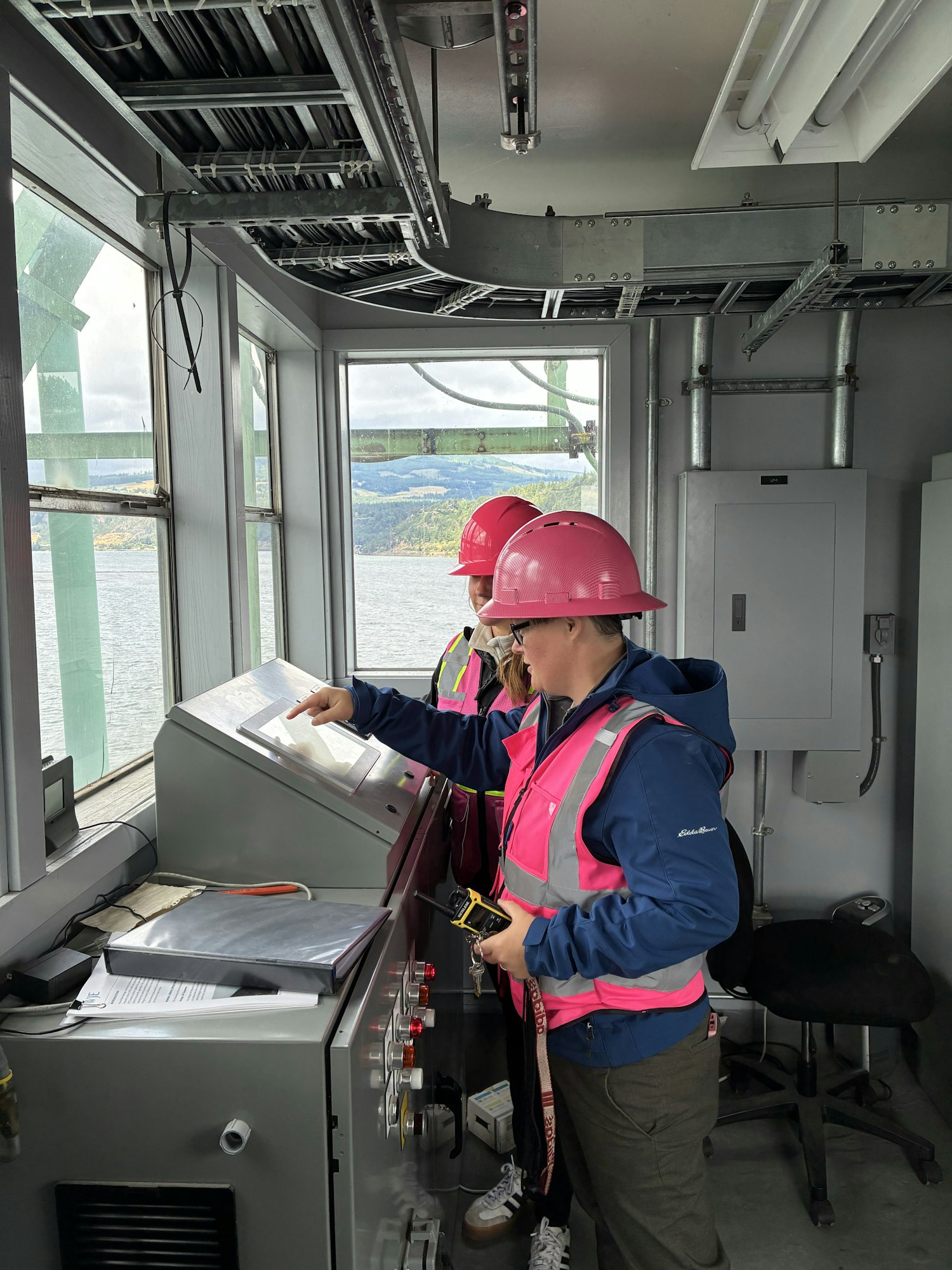 Two workers in pink safety vests and hard hats are inspecting equipment in a control room overlooking a body of water.