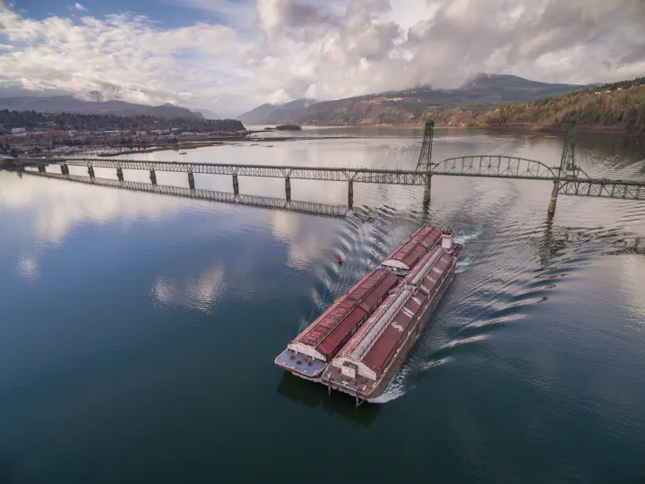A barge traveling under a metal bridge on a calm river surrounded by mountains.
