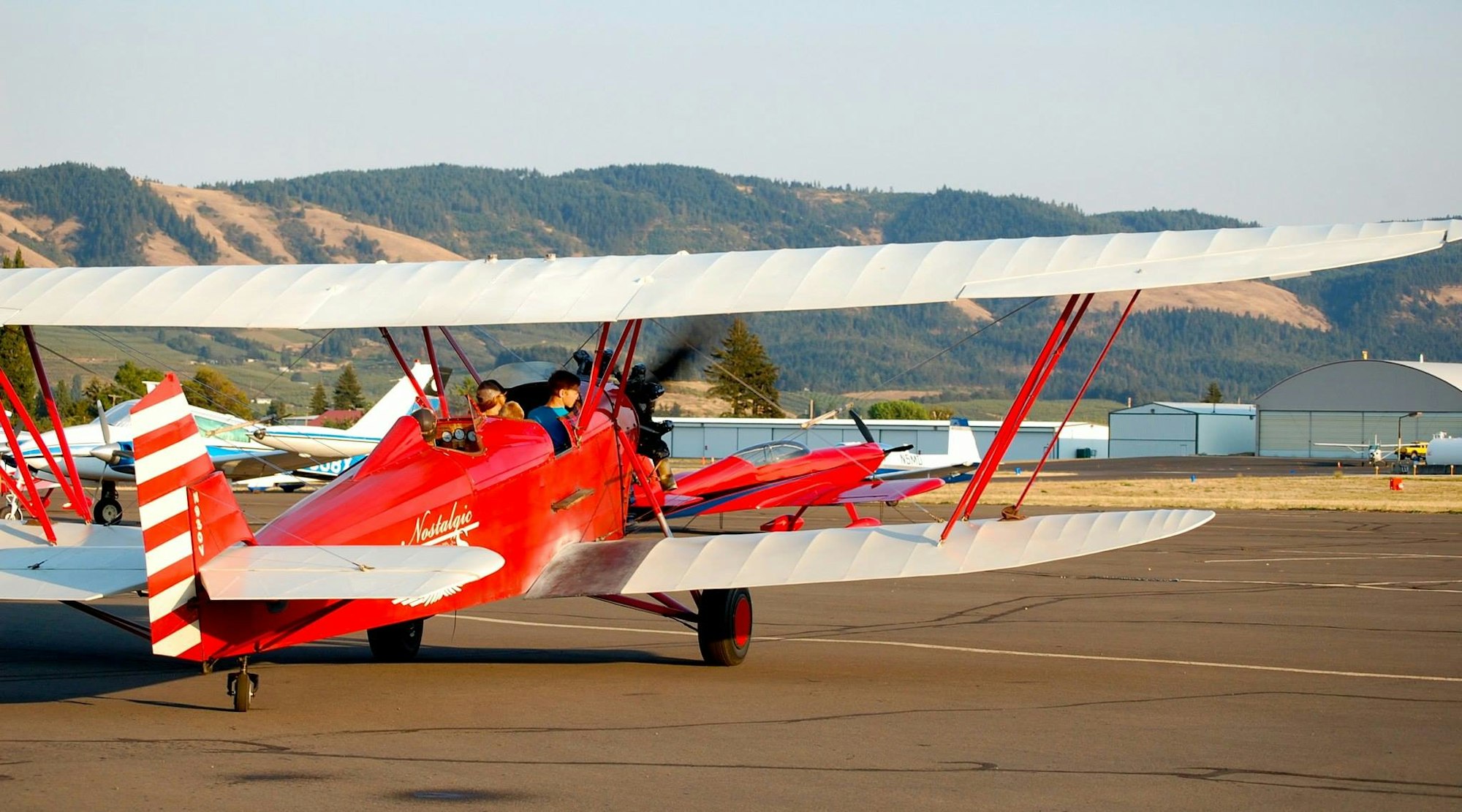 Red biplane on a runway, with passengers, against a backdrop of hangars and hills.
