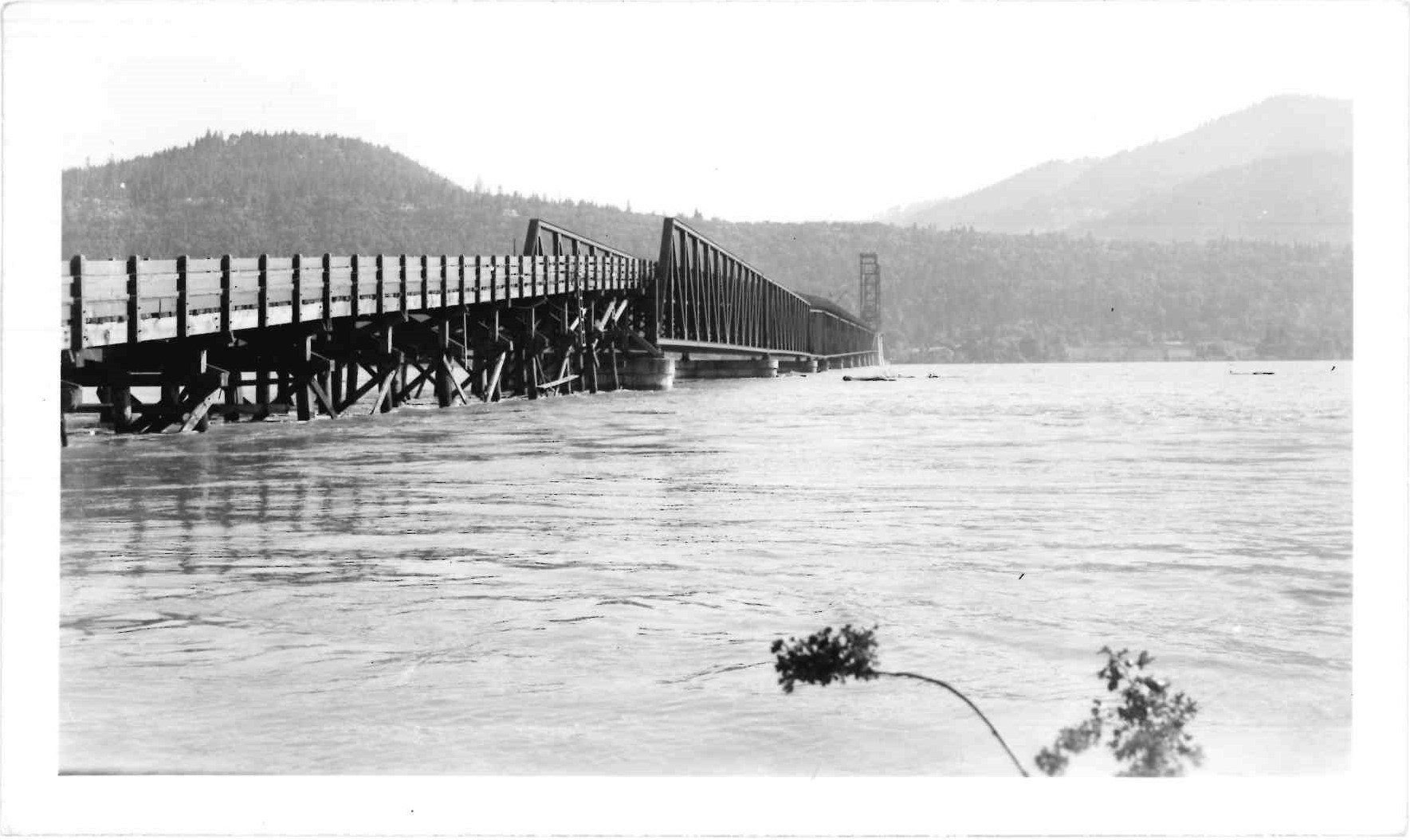 Historic black and white photo of a long wooden bridge over a river with hills in the background.