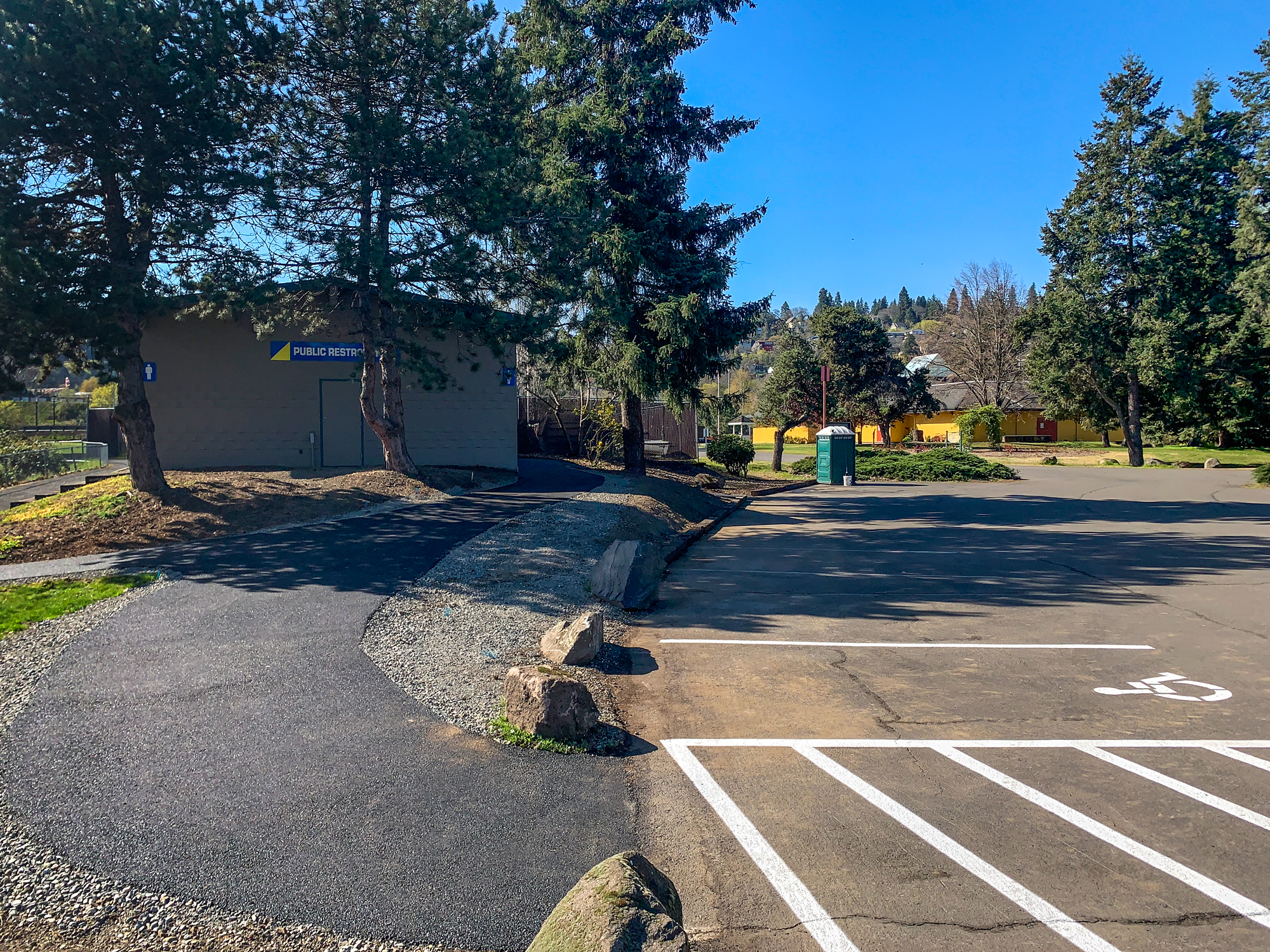 Public restroom building in a park with a clear sky, parking lot, and trees.