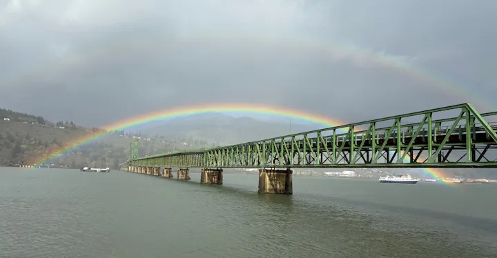 A green bridge stretches over a river with a vibrant rainbow arching above, set against a cloudy sky.