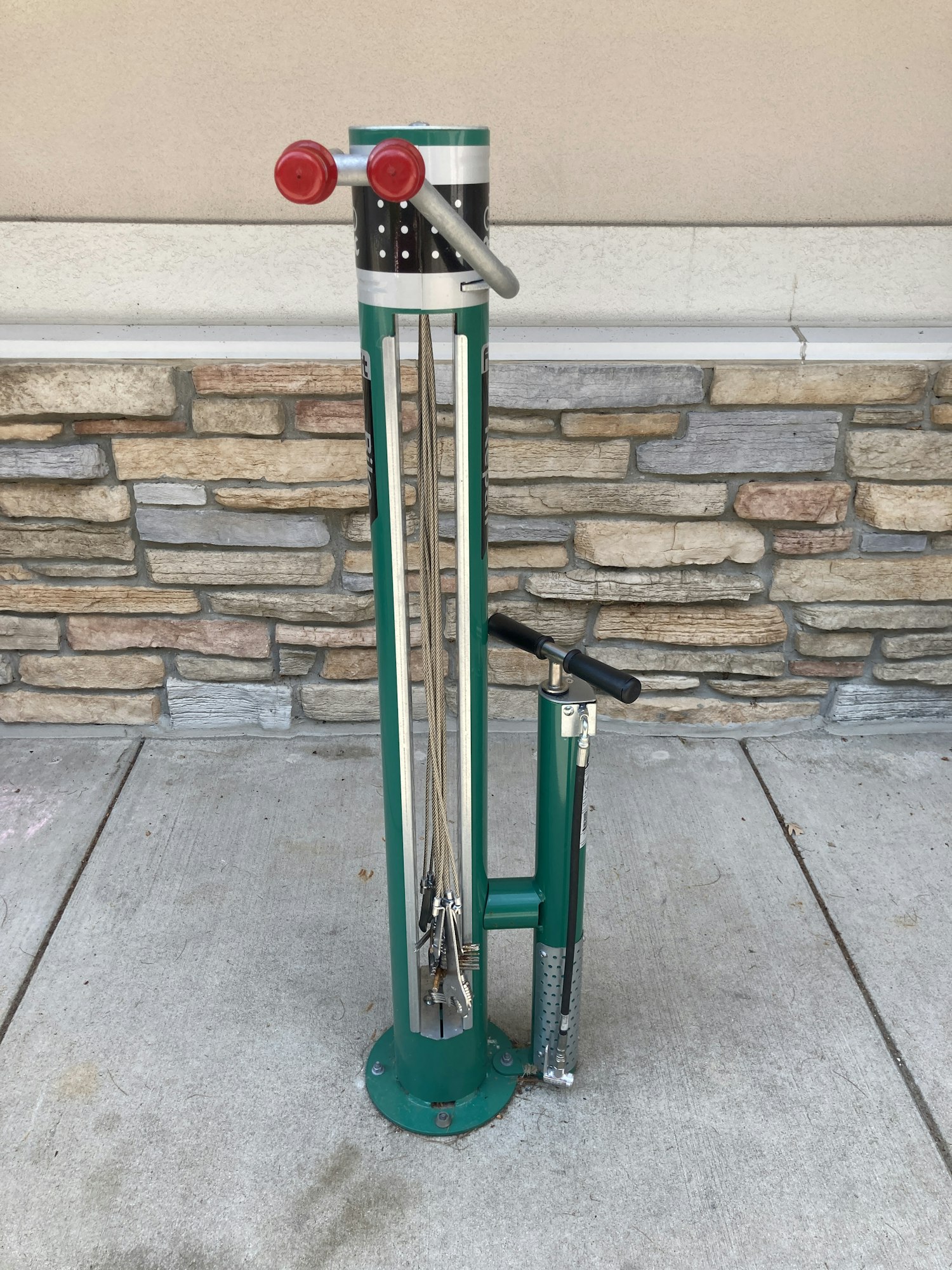 A green bike repair station with tools attached, mounted on a concrete sidewalk near a brick wall.