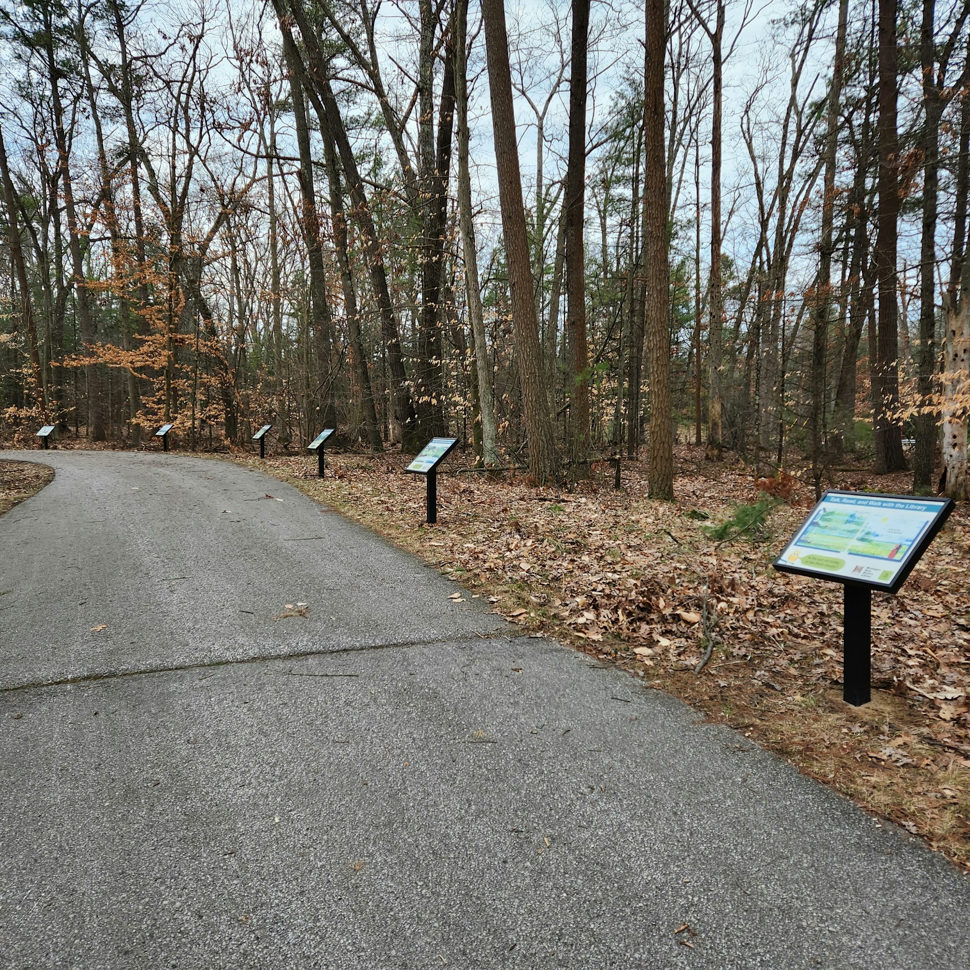 A wooded pathway with informational signs along the side, surrounded by trees and fallen leaves.