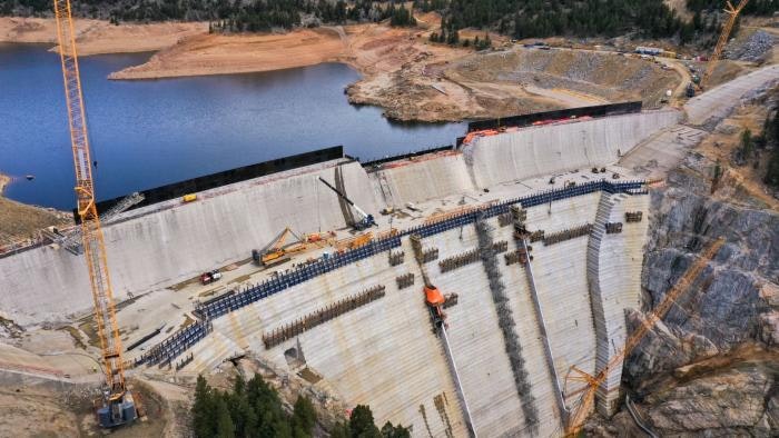 An aerial view of a large dam under construction, with heavy machinery and a reservoir in the background.