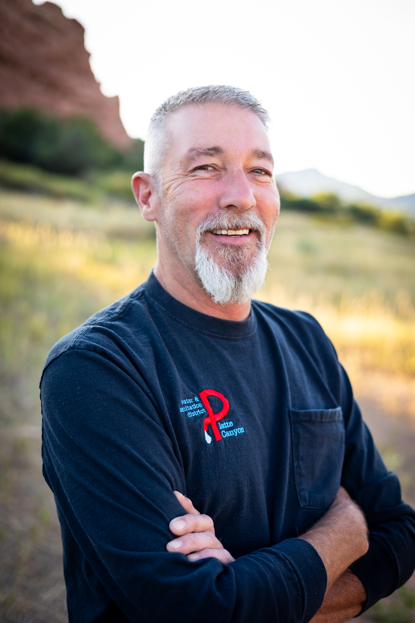 A smiling man with a beard stands outdoors, wearing a black shirt with a logo, surrounded by nature and mountains.