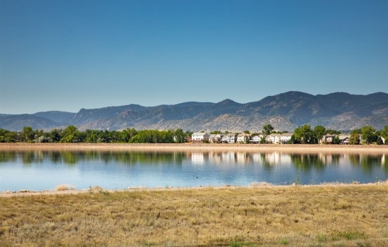 A serene landscape featuring a calm lake reflecting mountains and homes, with lush greenery and dry grass in the foreground.