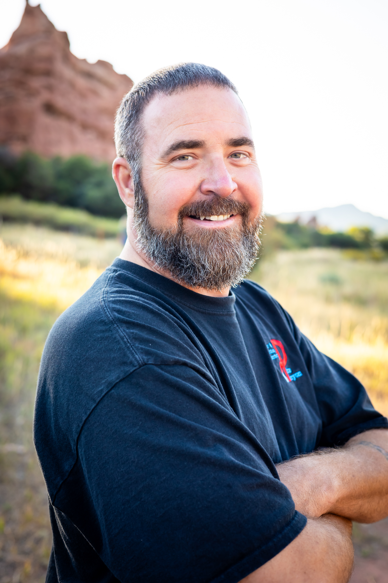 A smiling man with a beard stands with crossed arms, set against a beautiful natural background with rocks and greenery.