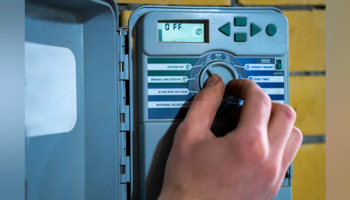 A person is adjusting a control panel for an irrigation system, showing options like "System Off" and "Manual-One Station."