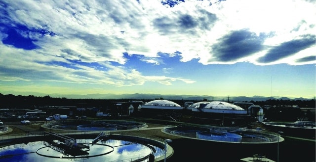 The image shows a landscape with circular water treatment tanks under a cloudy sky and mountains in the background.