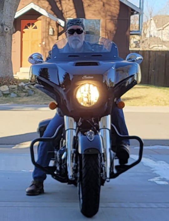 A person in sunglasses and a bandana is riding a black motorcycle in front of a house. Bright headlight is on.