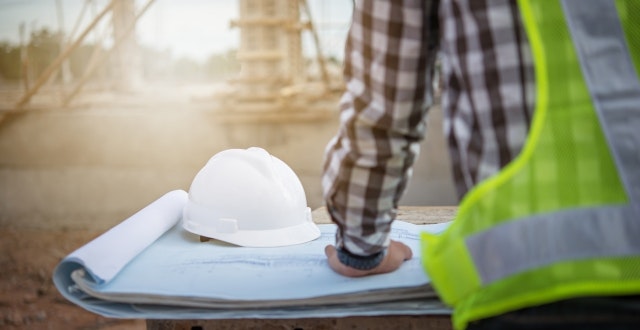 A construction worker examines blueprints on a site, with a hard hat nearby amidst a dusty background.