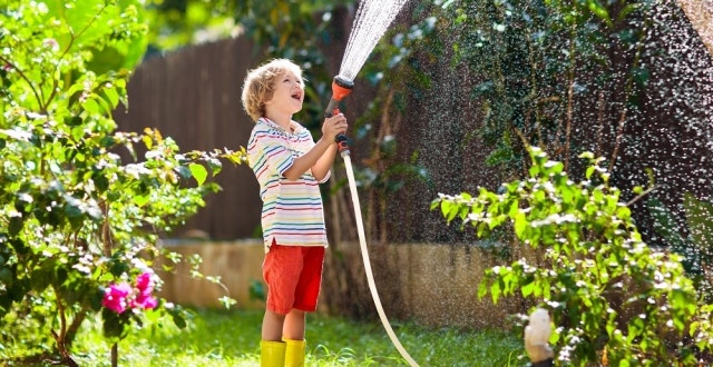 A joyful child in a garden, wearing a striped shirt and yellow boots, sprays water with a hose amidst blooming plants.