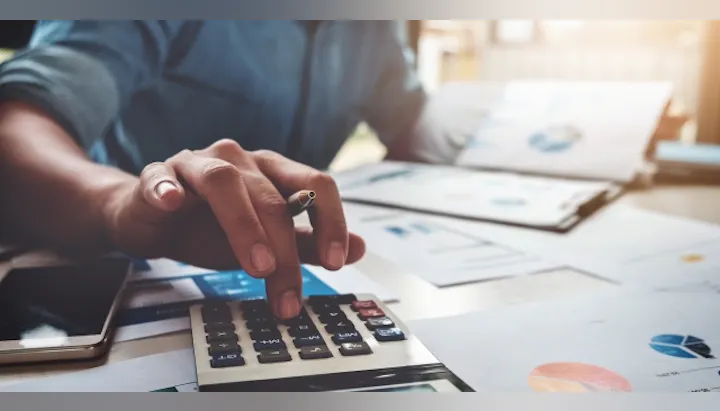 A person is using a calculator with one hand, while holding a cigarette and reviewing documents with charts and graphs on a desk.