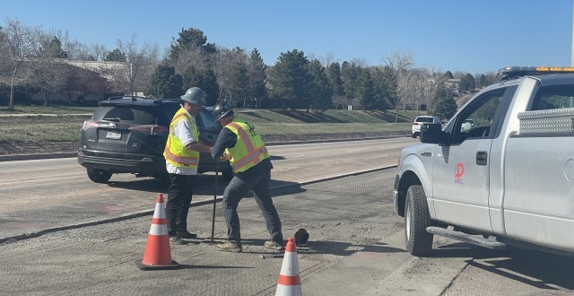 Two construction workers are working on a road, while a truck and traffic cones are present nearby.