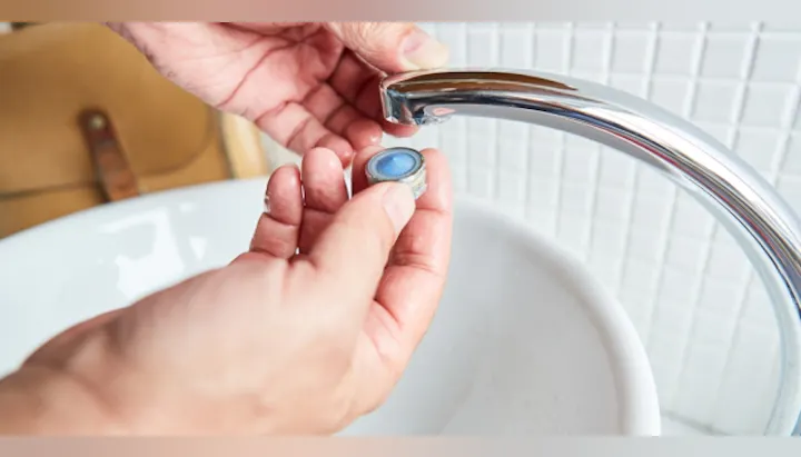 A person holds a small round object near a faucet above a sink, possibly for installation or repair purposes.