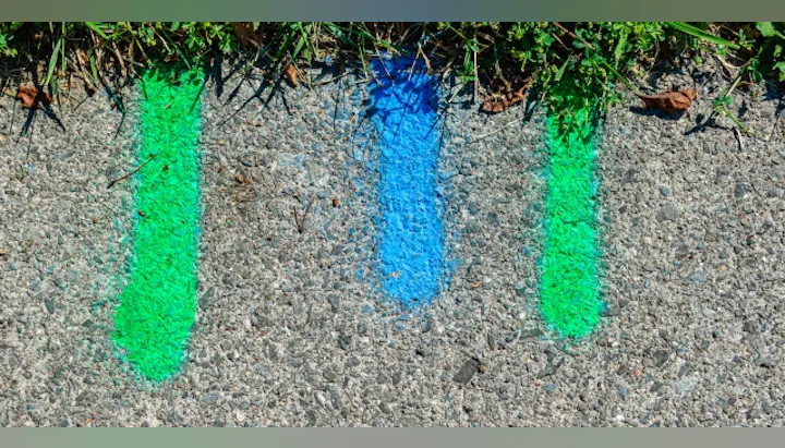 Three vertical lines of paint in green and blue on a concrete surface, surrounded by grass and a few leaves.