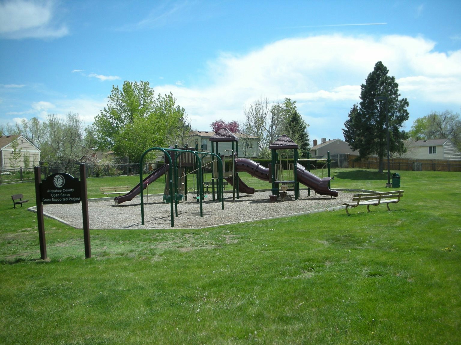 A playground with slides and climbing structures, a bench, and green grass with trees and houses in the background.