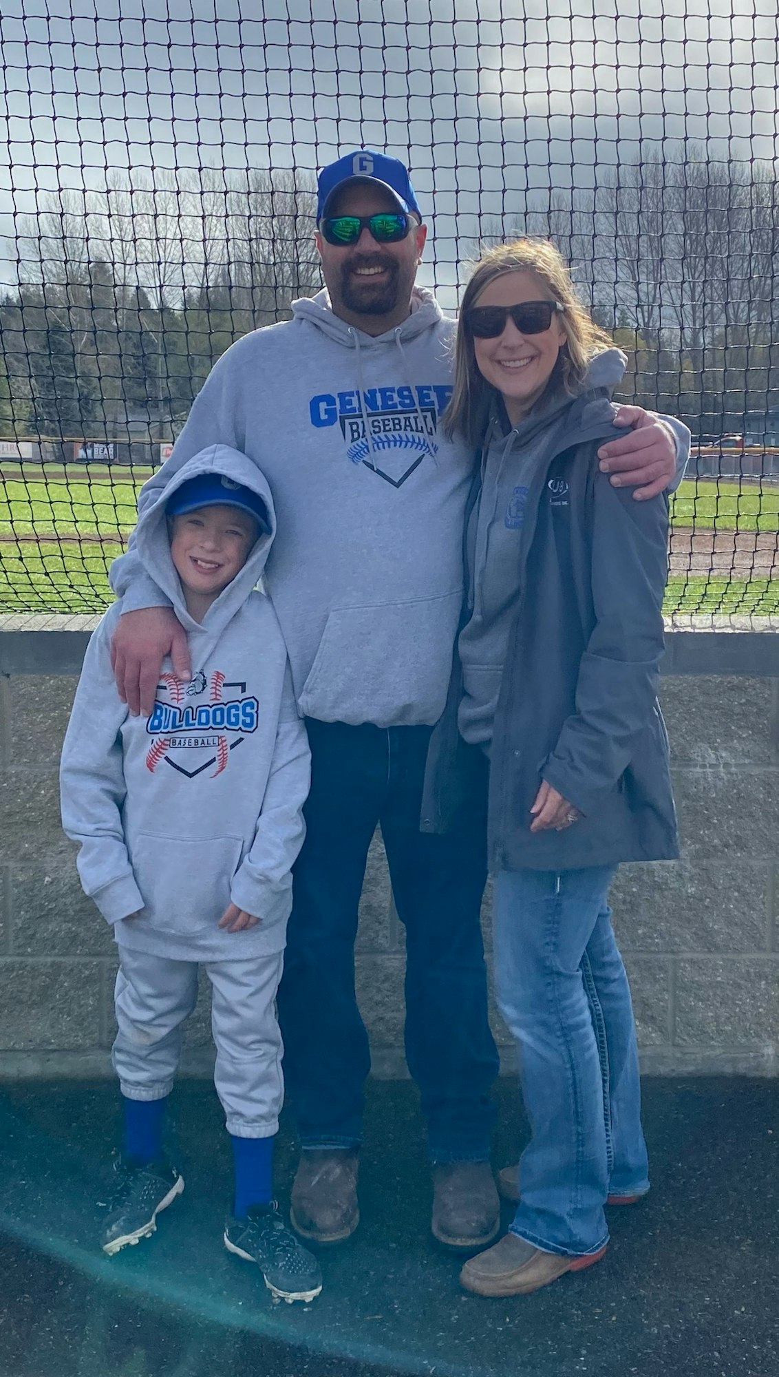 Three people smiling, wearing casual clothing with sports logos, standing behind a fence at what seems to be a sports field.