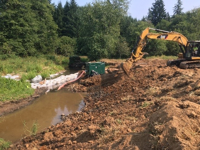 Excavator at work in a muddy construction site near water with trees in the background.