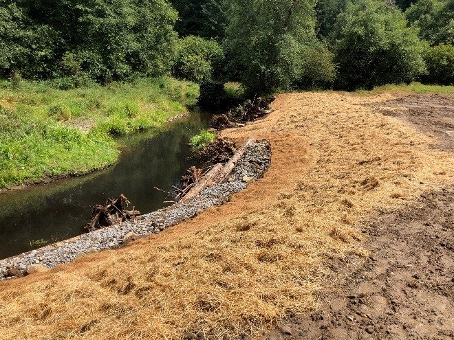 A stream, bank stabilization with rocks and logs, and a straw-covered slope.