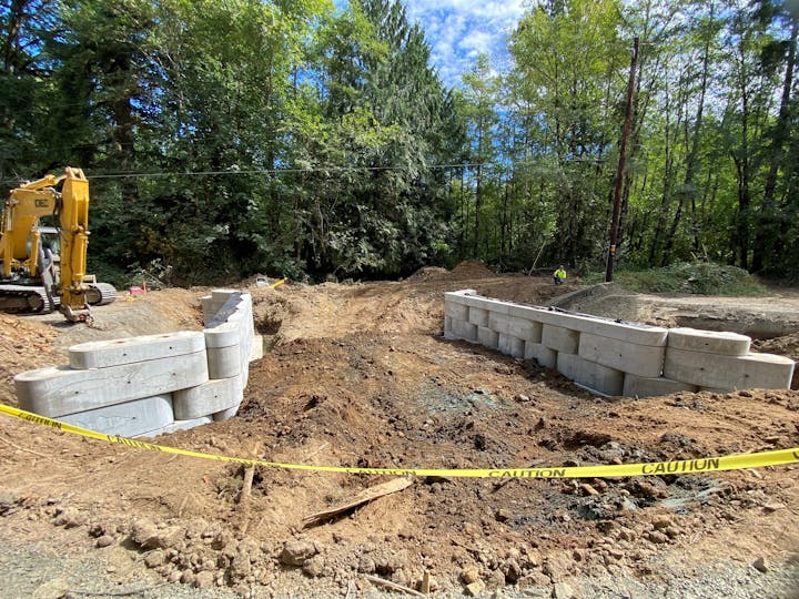 Construction site with large concrete blocks, an excavator, caution tape, and dense trees in the background.