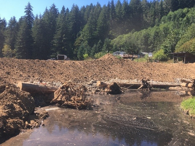 A creek with muddy banks, disrupted earth, trees, and infrastructure in the background.
