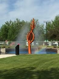 An orange sculpture resembling a flame with mist around it, set in a park with green grass and trees in the background.