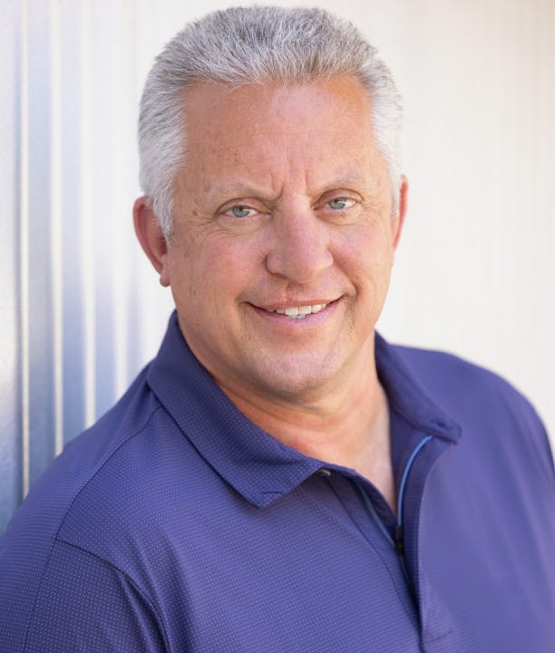 A man with gray hair smiles in a blue polo shirt against a light background.