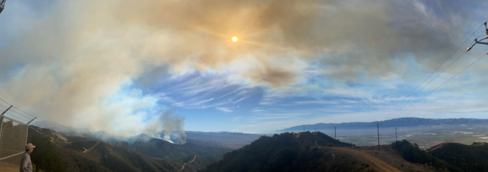 Panoramic view of a landscape showing smoke from a fire, with hazy sky and a person observing in the foreground.