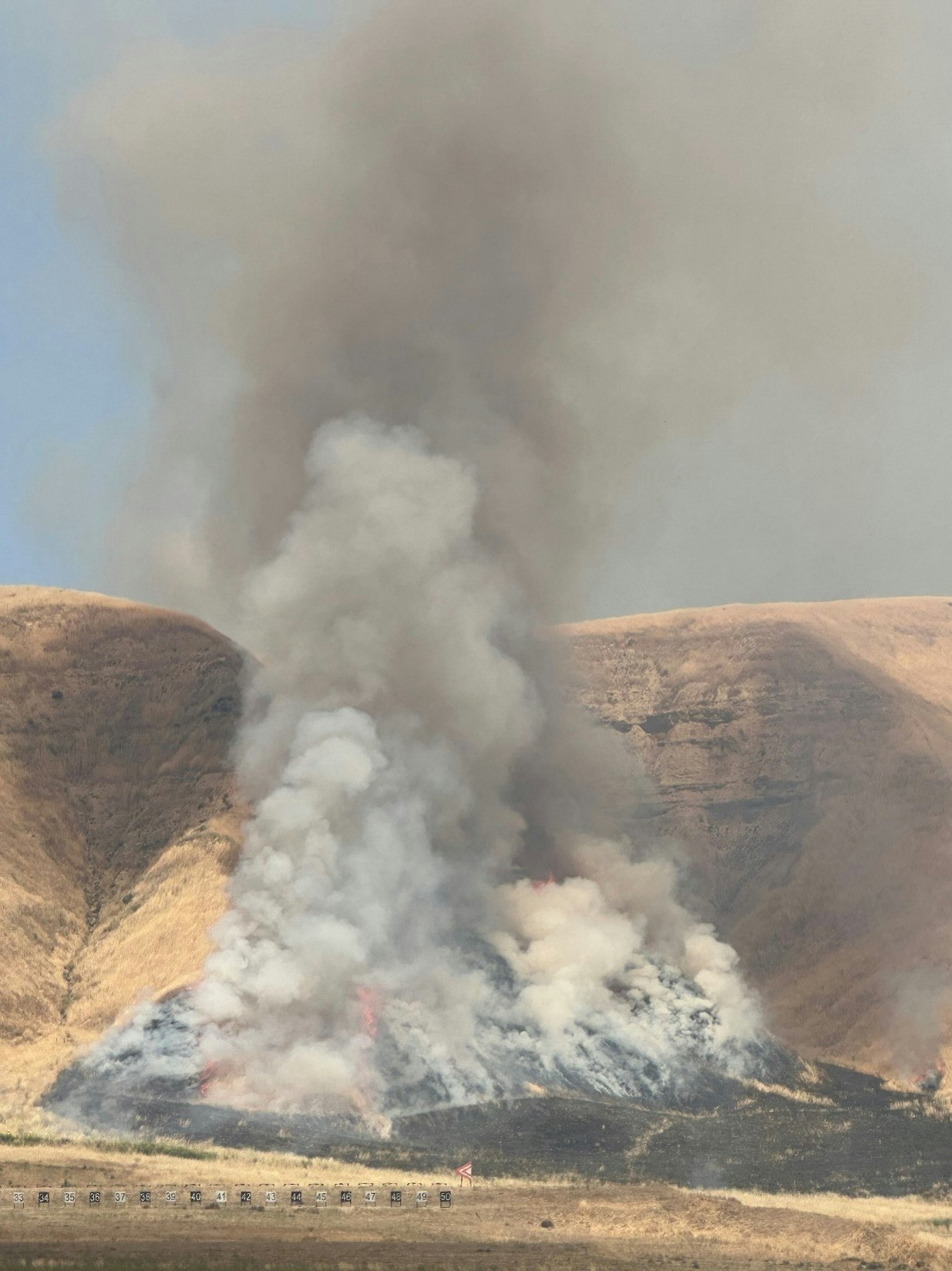 A wildfire is burning with smoke billowing, and dry hills in the background.