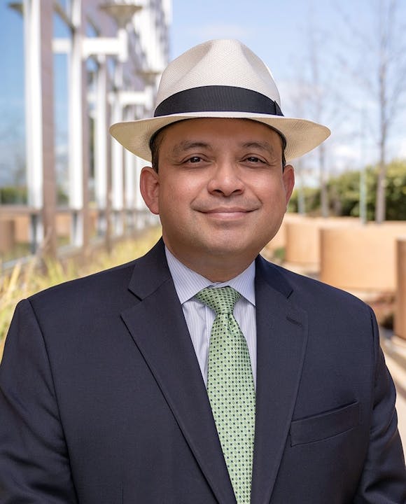 A man wearing a white fedora, suit, and green tie stands outdoors near a building.
