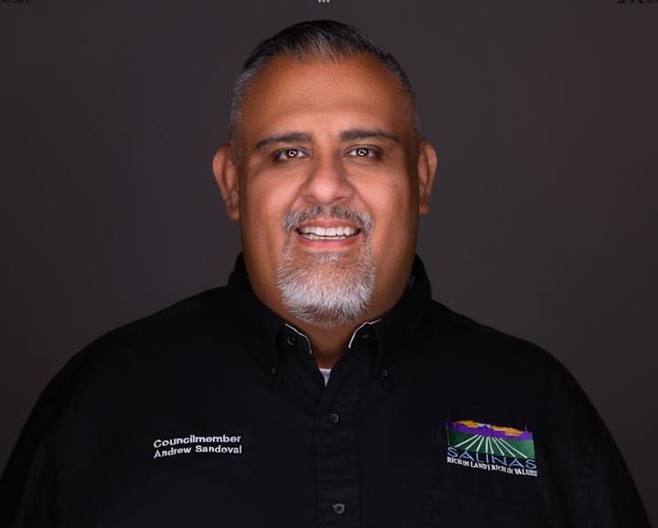 A person smiling, wearing a black shirt with logos and the name "Andrew Sandoval," identified as a councilmember.