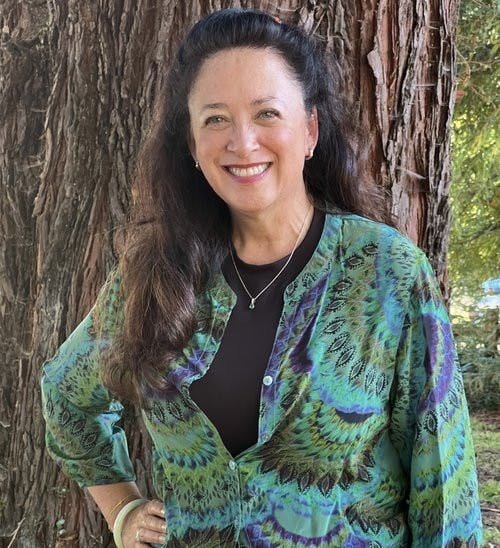 A woman smiling, standing in front of a tree, wearing a colorful top.