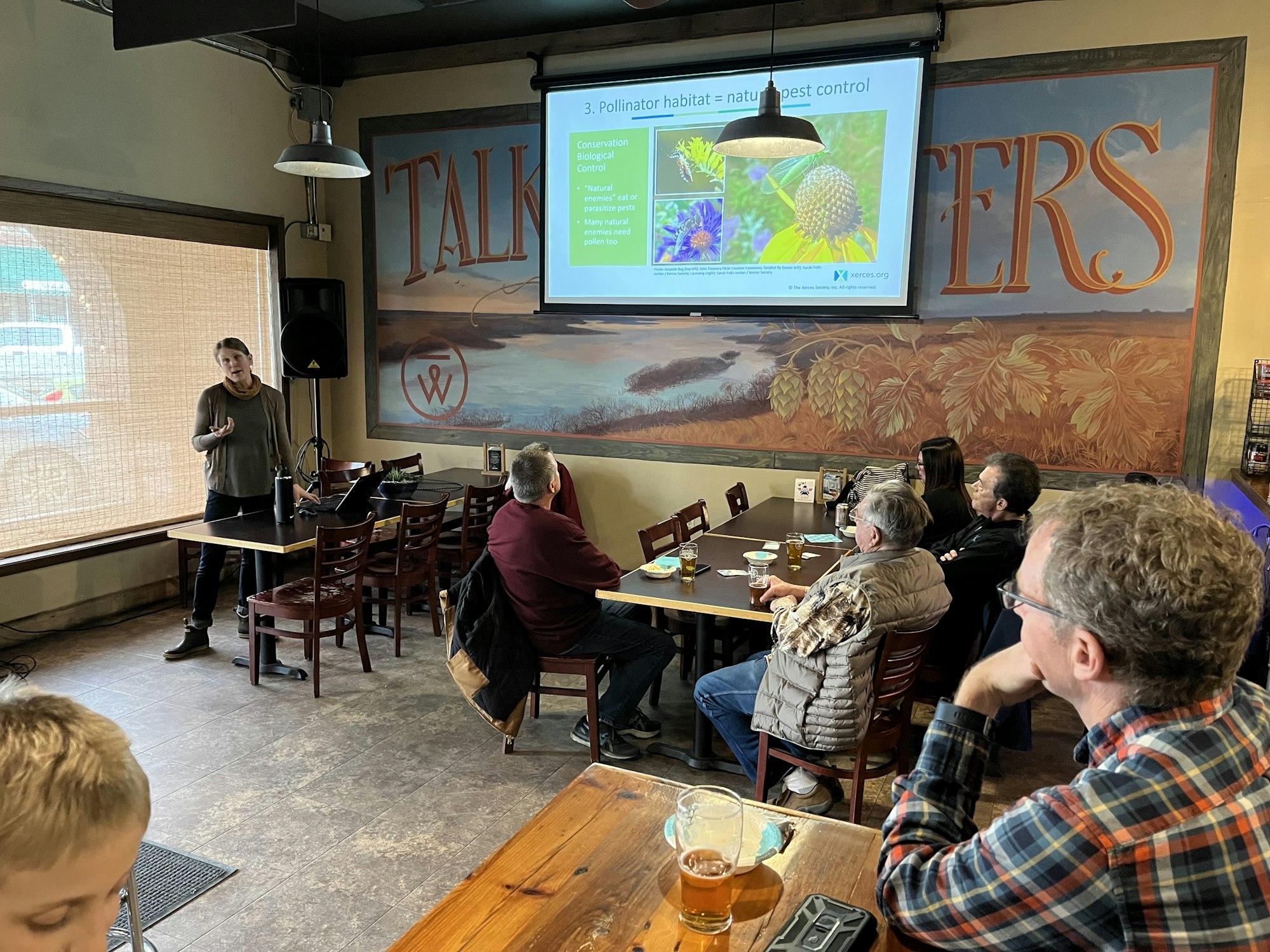 A person giving a presentation about pollinator habitats to a small group in a room with a mural and a projector screen.