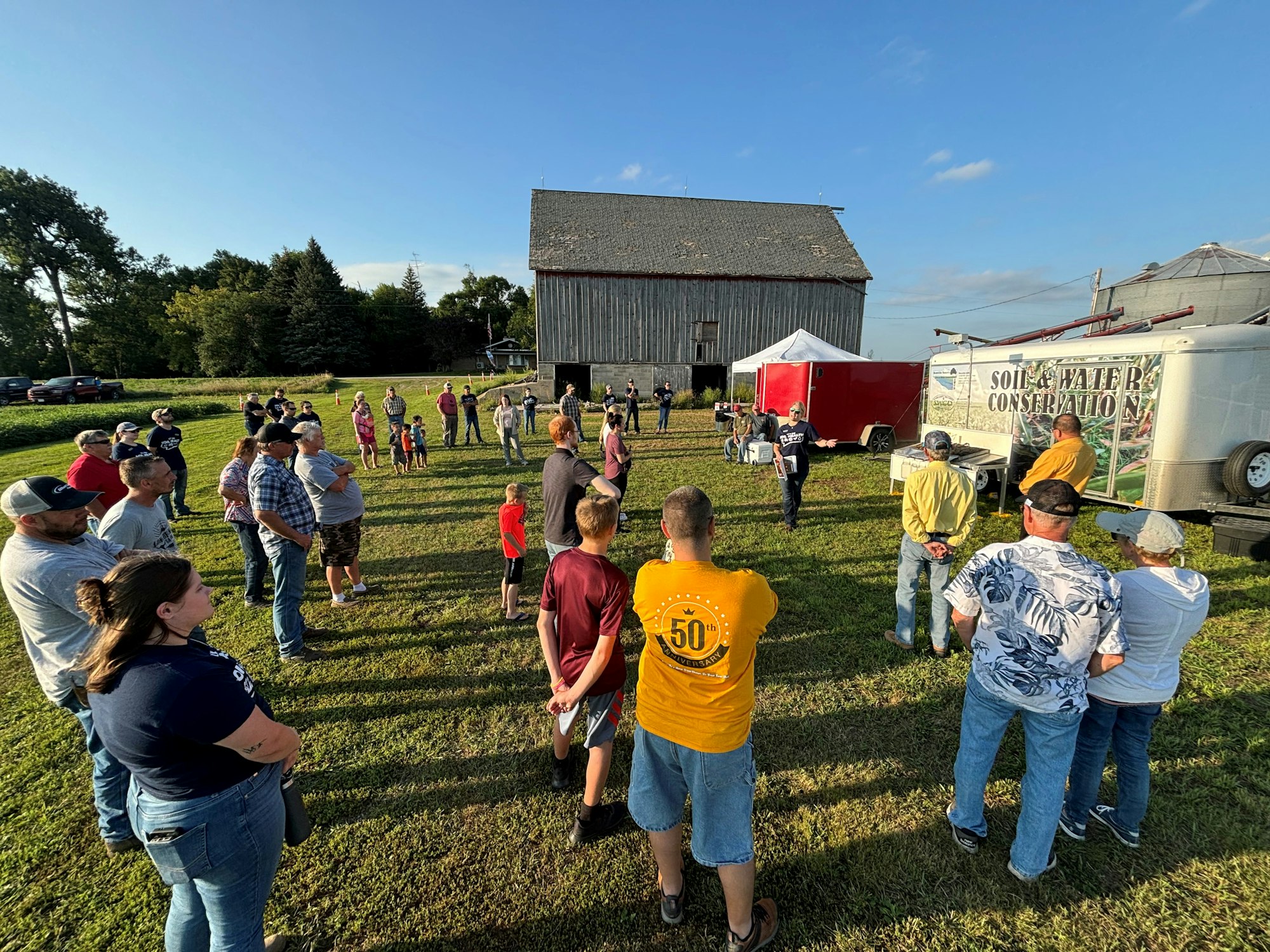 A group of people gather outside near a trailer marked "Soil & Water Conservation," by a barn under a clear blue sky.