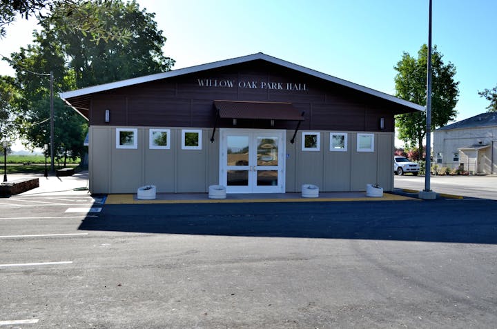 A brown building labeled "Willow Oak Park Hall" with a glass door entrance, flanked by windows and trees, in a parking lot.