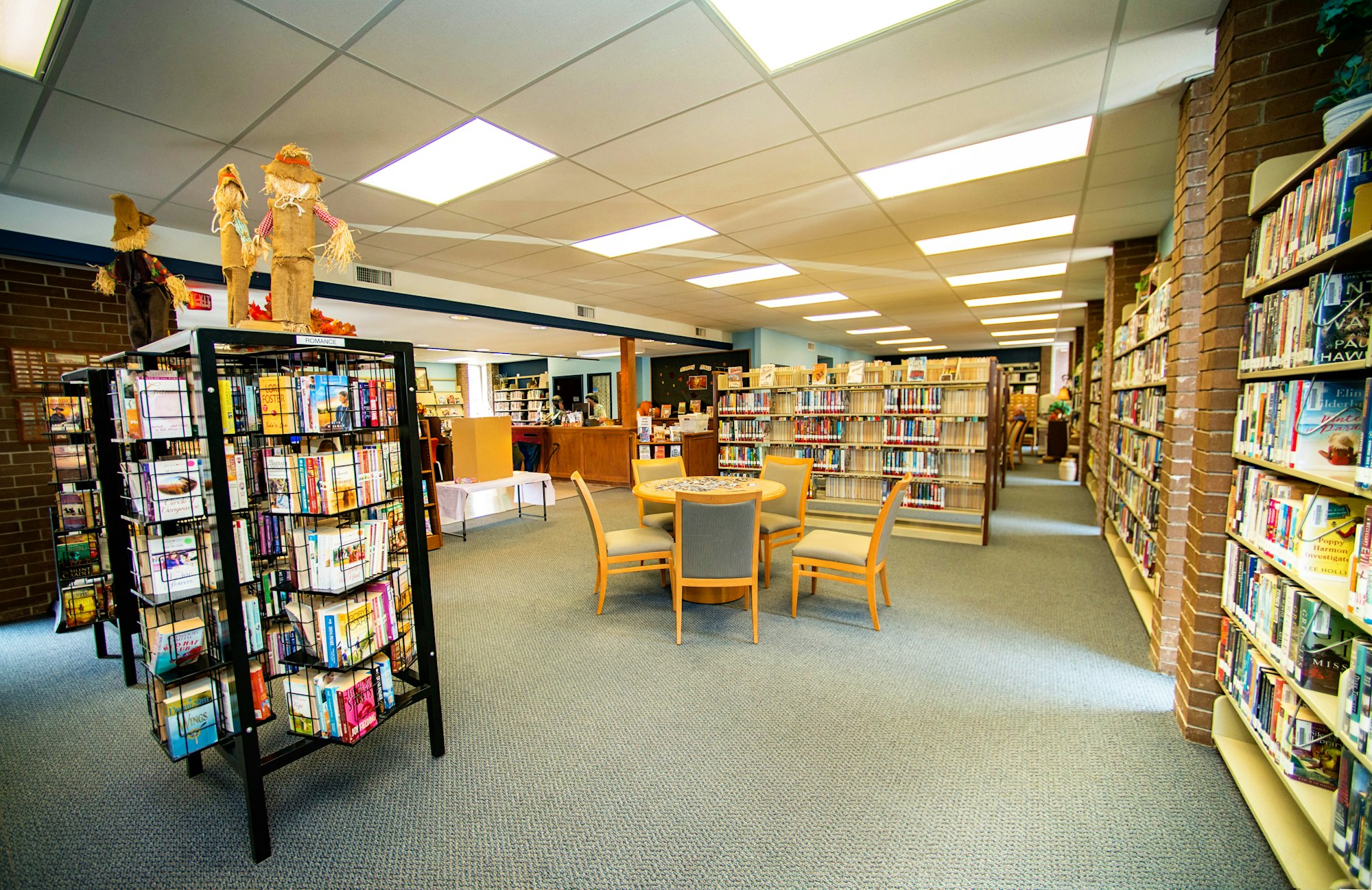 A bright library interior with bookshelves, a rotating rack of books, seating area, and scarecrow decorations.