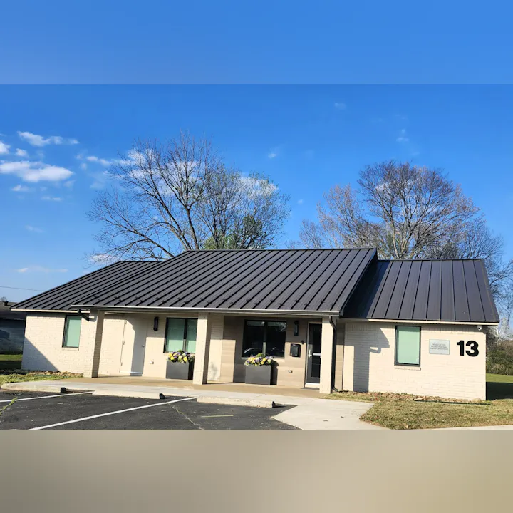 A modern building with a metal roof, featuring large windows and flower planters, set against a clear blue sky.