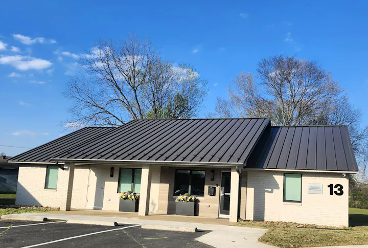 A modern building with a metal roof, featuring large windows and flower planters, set against a clear blue sky.