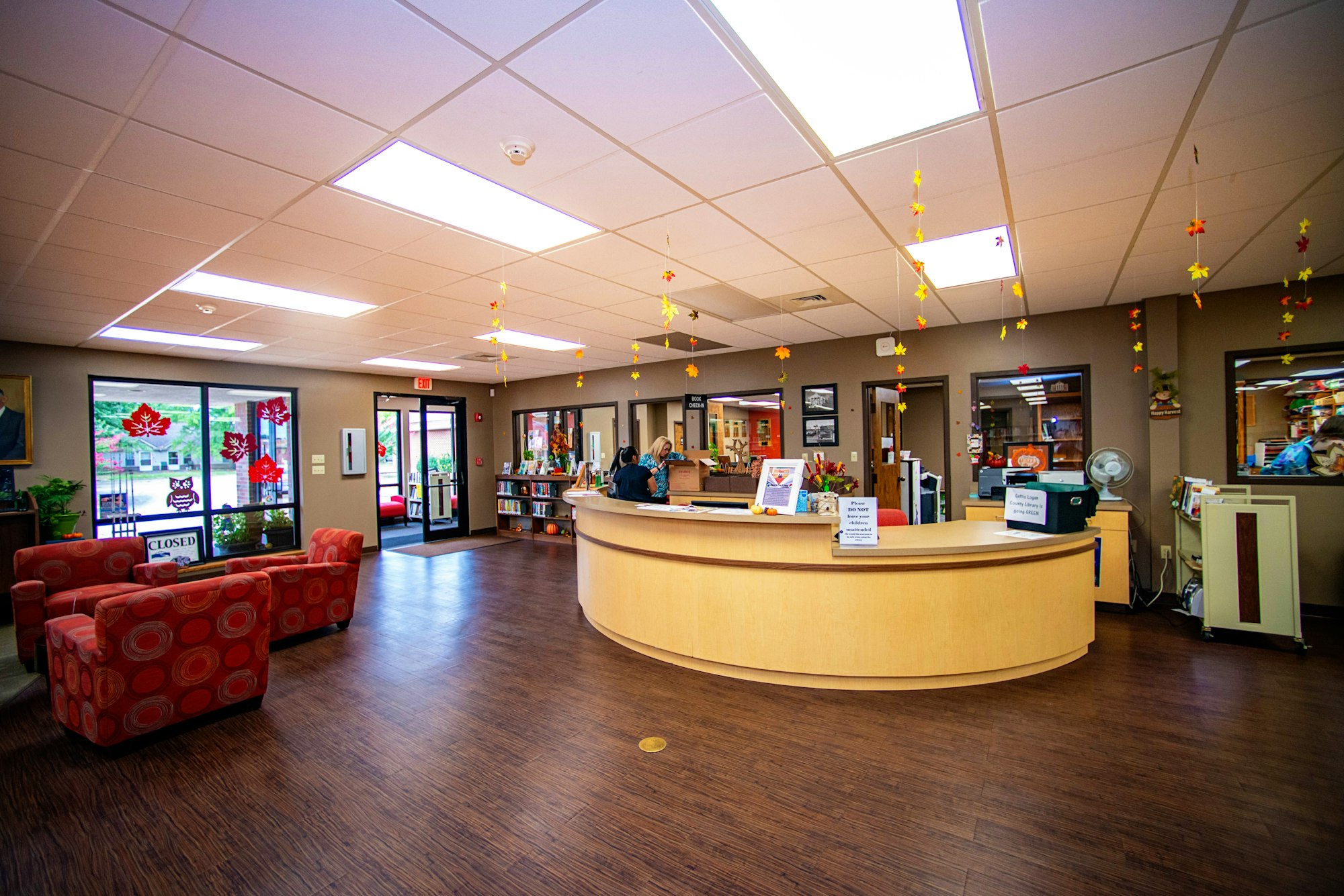 An inviting, well-lit lobby with a circular reception desk, cozy seating area, fall decorations, and a view into a book-filled space.