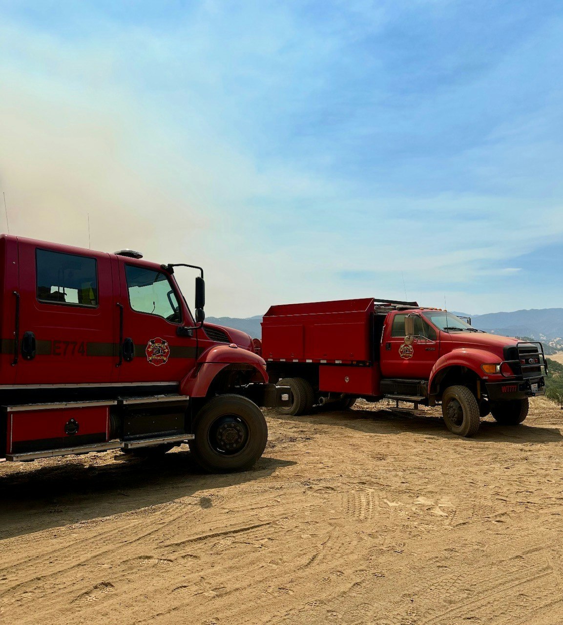 Two red trucks parked on a dirt road, likely used for firefighting, with mountains and a smoky sky in the background.