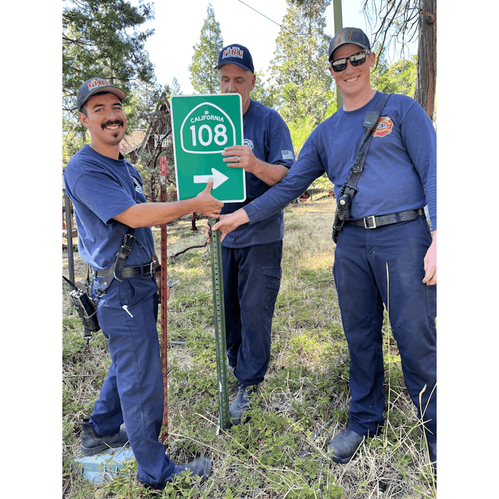 Three people in uniforms installing a California Route 108 road sign outdoors.