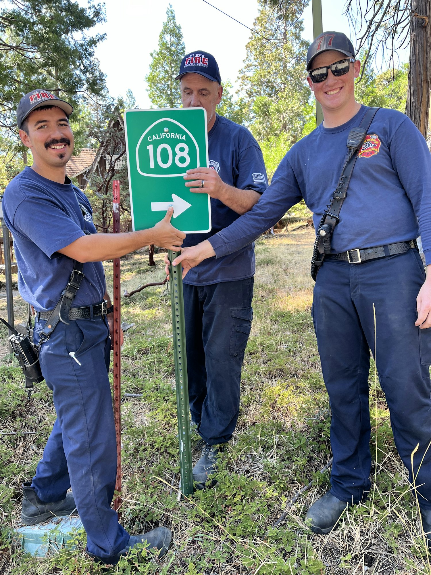Three people in uniforms installing a California Route 108 road sign outdoors.