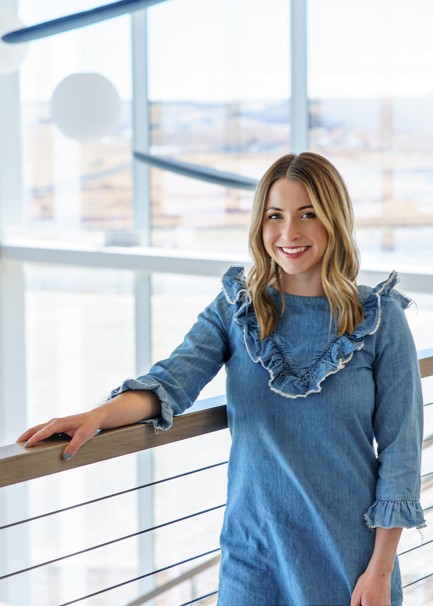 A person in a denim dress, smiling, leaning on a railing in a bright, modern space.
